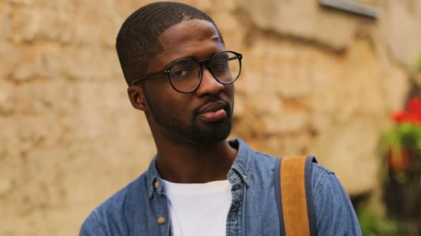 Retrato de atraente jovem africano em óculos posando para a câmera e sorrindo no fundo da cidade. Ao ar livre . — Vídeo de Stock
