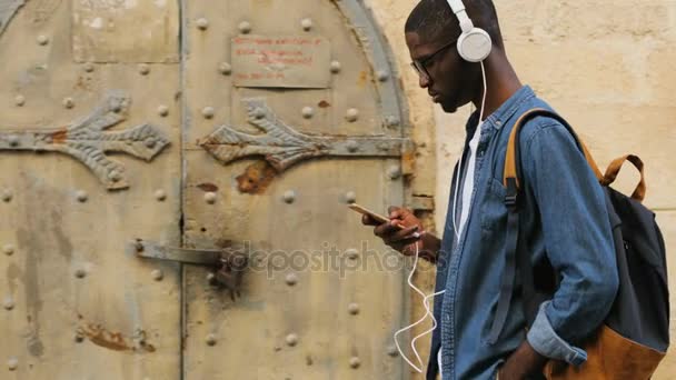 Seitenansicht eines jungen Afrikaners mit Brille, der Musik über Kopfhörer und Smartphone in der Stadt hört. Außenbereich. — Stockvideo