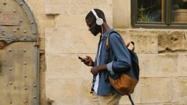 Vista lateral del guapo turista africano en gafas y auriculares escuchando música usando un teléfono inteligente que va a la ciudad . — Vídeos de Stock
