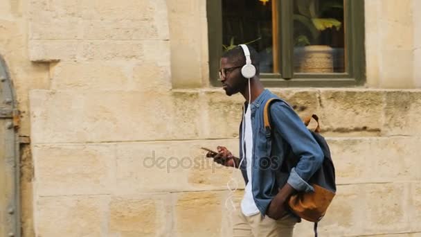 Zijaanzicht van aantrekkelijke Afrikaanse mannelijke toeristische in glazen luisteren naar muziek met behulp van hoofdtelefoons en slimme telefoon gaan in de stad. — Stockvideo
