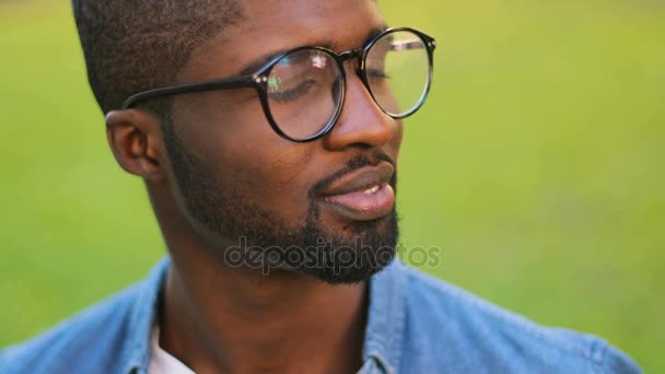 Portrét přátelské africké muž s brýlemi v modré košili pózuje pro kameru, usmívá se zuby a ukazující Ok. Venkovní. — Stock video