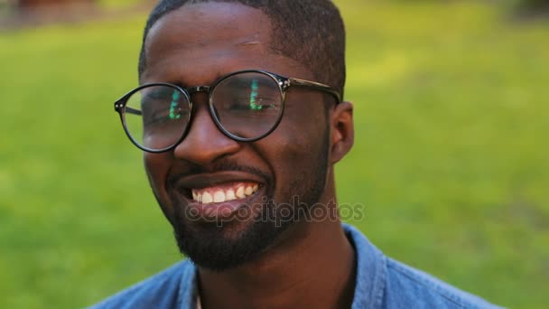 Porträtt av attraktiva vänliga afrikanska mannen med glasögon i blå skjorta poserar för kameran och ler med tänder. Utomhus. — Stockvideo