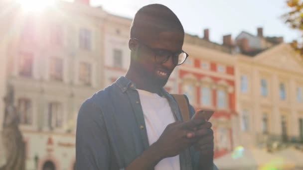 Bonito joven africano con gafas en camisa azul utilizando el teléfono móvil de pie en el centro de la ciudad. Exterior . — Vídeos de Stock