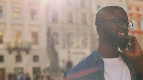 Ritratto di attraente uomo africano in camicia blu che parla su smartphone con amico e sorride con i denti in città. All'aperto . — Video Stock