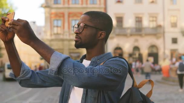 Молодих афро-американських туристів в окулярах в блакитній сорочці, за допомогою мобільного телефону за прийняття фотографії, що стоїть в центрі міста. — стокове відео