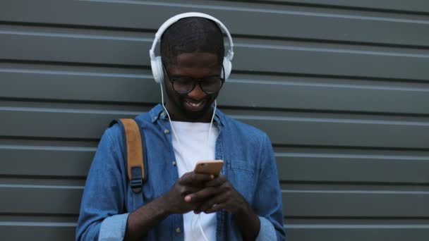 Atractivo turista afroamericano joven en auriculares charlando con un amigo usando un teléfono inteligente. Exterior . — Vídeos de Stock
