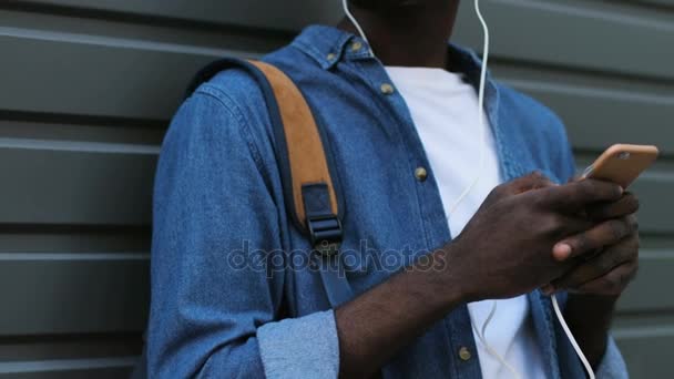 Pohledný mladý africké americký turista poslechu hudby pomocí bílé sluchátka a black chytrý telefon. Venkovní. — Stock video