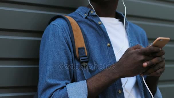 Handsome young african american tourist listening to music using white headphones and black smart phone. Outdoor. — Stock Video