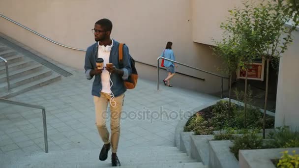 Giovane uomo africano in occhiali con le cuffie utilizzando smartphone e tenendo il caffè in mano mentre va sulle stelle in città . — Video Stock