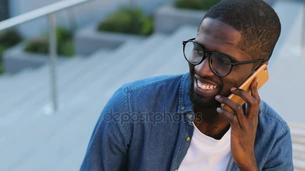 Portrait of handsome african man talking on mobile phone and drinking coffee while sitting on the bench on background with stairs. — Stock Video