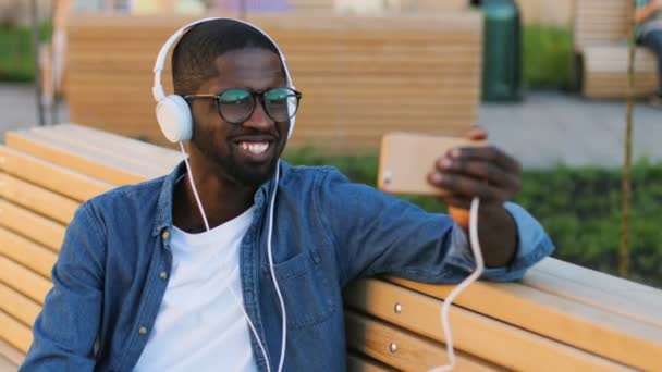Homem africano atraente em óculos com fones de ouvido usando telefone inteligente para vídeo conversando com amigo localizado no banco da cidade . — Vídeo de Stock