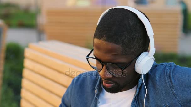 Portret młodzieńca Afryki w okularach zdejmowania słuchawek, patrząc na kamery i uśmiechnięty, siedzi na ławce w mieście. — Wideo stockowe
