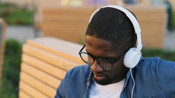 Portrait of young african man in glasses removing headphones, looking to the camera and smiling, siting on the bench in city. — Stock Video