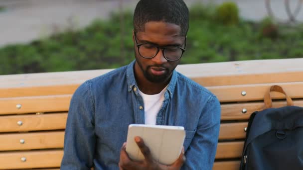 英俊的非洲年轻人中使用的眼镜片选址在公园的长凳上，看不到相机，微笑. — 图库视频影像