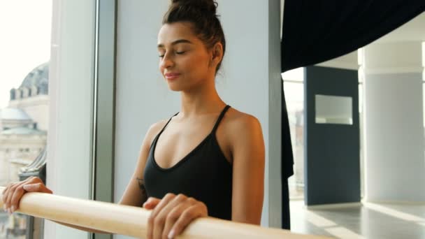 Portret atrakcyjny brunetka baleriny stojący w pobliżu balet barre w studio i uśmiecha się na aparat, landskype miasto z bliska. — Wideo stockowe