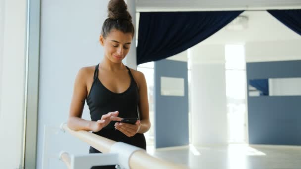 Gros plan portrait d'une jolie ballerine brune debout près de la barre de ballet et utilisant un téléphone intelligent . — Video