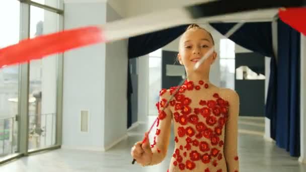 Портрет маленькой изящной гимнастики, вращающей красную ленту перед камерой в белой студии . — стоковое видео