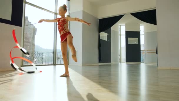 Pequeña gimnasia en cuerpo rojo haciendo el ejercicio de gimnasia con una cinta en el elegante estudio y posando a la cámara. Ver desde la parte inferior . — Vídeo de stock