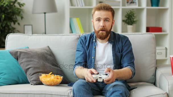Unge känslomässiga mannen med skägg som spelar spel på xbox medan du sitter på soffan hemma i vardagsrummet. — Stockvideo