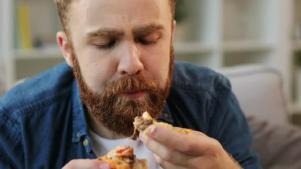 Portret van jonge hipster met baard die het eten van pizza op home op de achtergrond van de woonkamer. Close-up. — Stockvideo