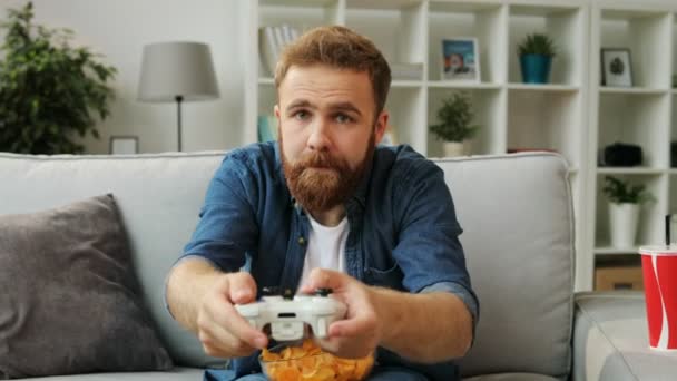 Pemuda emosional bermain video game di xbox, makan keripik sambil duduk di sofa di ruang tamu . — Stok Video