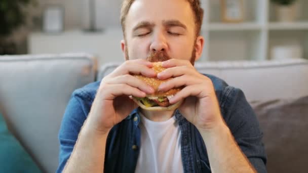 Portret młodzieńca jedzenia fast foodów humburger przed kamerą na tle pokój dzienny. Z bliska. — Wideo stockowe