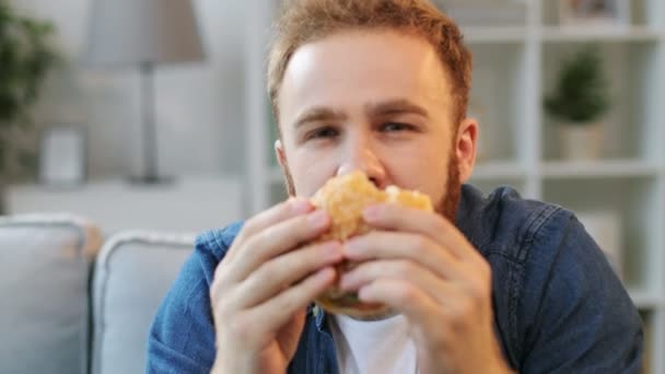 Portrét mladého muže jíst nezdravé jídlo humburger, zatímco sedí na gauči doma v obývacím pokoji a sledovat zajímavý film. Detailní záběr. — Stock video