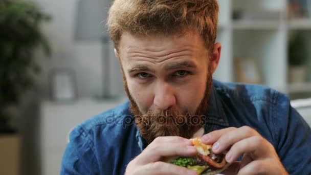 Portrait of young man eating junk food humburger and teasing it in front of camera on living room background. Close up. — Stock Video