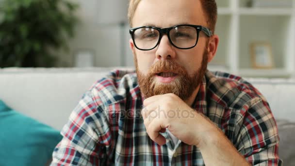 Portret młodego człowieka poważne w kubki siedzi przed kamerą, oglądając film w domu w salonie z bliska. — Wideo stockowe