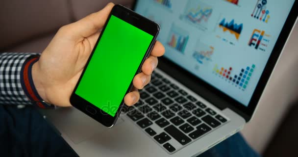 Κοντινό πλάνο άνθρωπος χέρια κρατώντας έξυπνο τηλέφωνο με πράσινη οθόνη. Κλειδί Chroma. Κοντινό πλάνο αρσενικό χέρια κύλιση σελίδες, εικόνες στο κινητό με φόντο το laptop. Επαγγελματικά γραφικά — Αρχείο Βίντεο
