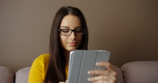 Красивая женщина в очках с помощью планшетного устройства сидит на диване в гостиной. Молодая женщина печатает на сенсорном экране и улыбается — стоковое видео