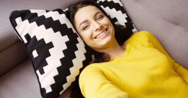 Retrato de una hermosa joven en suéter amarillo acostada en la almohada en el sofá de la sala de estar. Retrato de mujer atractiva sonriendo a la cámara. Primer plano. — Vídeos de Stock