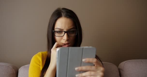 Bella donna in bicchieri utilizzando dispositivo tablet seduto sul divano in soggiorno. Giovane donna digitando sul touch screen e sorridente — Video Stock