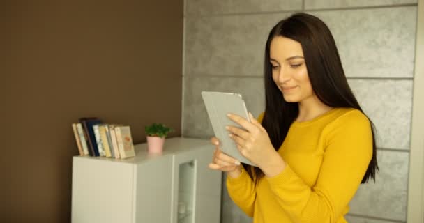 Donna attraente digitando sul dispositivo tablet touchscreen e sorridendo a casa. Bella giovane donna che utilizza il suo computer tablet al coperto . — Video Stock