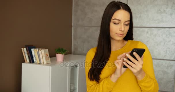 Hermosa mujer en suéter amarillo que sostiene el teléfono celular en el interior. Mujer joven usando el teléfono inteligente, escribiendo en la pantalla y navegar por Internet en casa . — Vídeo de stock