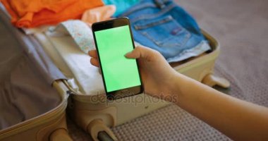 Akıllı telefon tutarak ve yeşil ekran sayfalarla kaydırma kadın el kapatın. Seyahat çanta, bavul arka plan. Preparing için a çelme takmak, tatil. Seyahat. Chroma anahtar