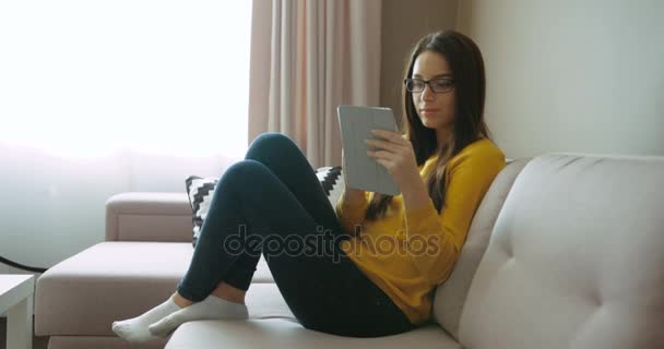 Hermosa mujer en gafas con dispositivo de tableta mientras se sienta en el sofá en la sala de estar. Mujer joven escribiendo en la pantalla táctil y sonriendo. Vista lateral — Vídeos de Stock