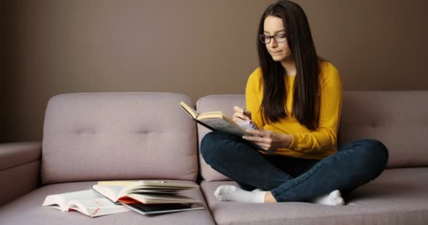 Menina estudante bonita sentada no sofá, stying, lendo livros, observando e fazendo lição de casa em casa . — Vídeo de Stock