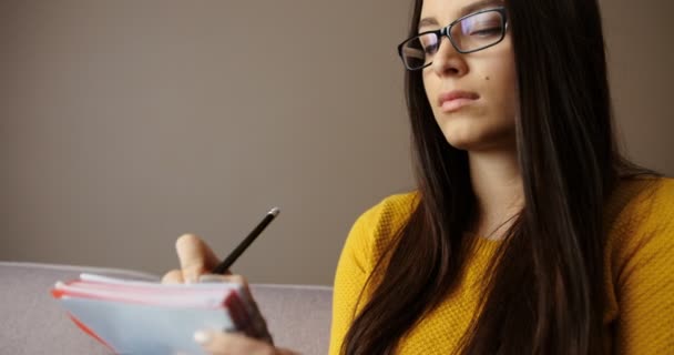 Close-up shot van mooie jonge vrouw in glazen zittend op de Bank, vaststellend in notitieblok, studeren, lezen van boeken in de woonkamer — Stockvideo