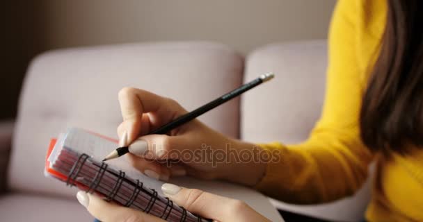 Крупним планом жіночі руки пишуть в блокноті. Жінка сидить на дивані у вітальні і відзначає. Близько. вид збоку — стокове відео