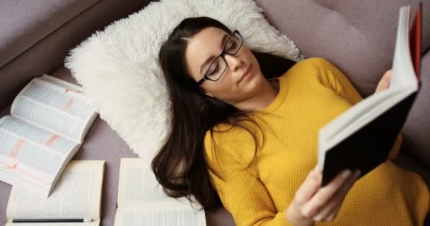 美丽的女人躺在沙发上和研究，在家里读书。黄色的毛衣和学习室内的眼镜的女人。顶视图 — 图库视频影像