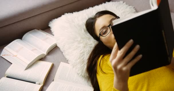 Nahaufnahme attraktive Frau, die auf dem Sofa liegt und studiert, Bücher zu Hause liest. Frau in gelbem Pullover und Brille, die drinnen studiert. Ansicht von oben. Schülerin — Stockvideo