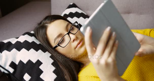Νεαρή γυναίκα σε τζάμια, ανάγνωση σε απευθείας σύνδεση βιβλίο στον υπολογιστή tablet ξαπλωμένοι στον καναπέ στο σαλόνι. — Αρχείο Βίντεο