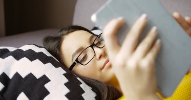 Attractive young woman using tablet device while lying on the couch. Beautiful woman in glasses typing on tablet computer and smilling at home. Close up — Stock Video