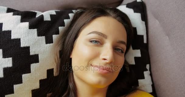 Portret van mooie vrouw die lacht naar de camera. Portret van mooie jonge vrouw in gele trui liggen in het kussen op de Bank in de woonkamer. Close-up shot — Stockvideo