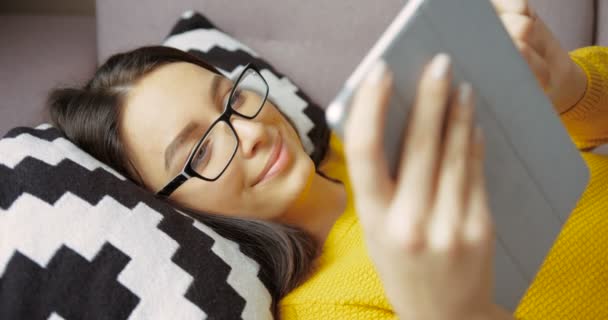 Девочка пользуется планшетным компьютером, стучит по сенсорному экрану, лежа на диване в гостиной. Улыбаясь в ожидании фильма на планшете — стоковое видео