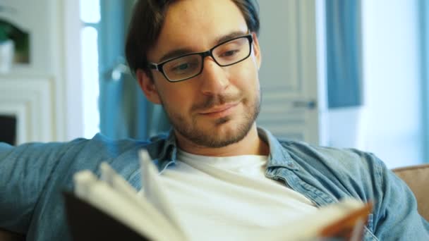 Jeune homme attrayant avec des lunettes de détente sur le canapé à la maison et le livre de lecture. Gros plan . — Video