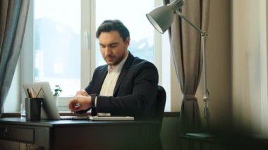 Genç ciddi iş adamı uygun office arka pencere ile dizüstü bilgisayar ve akıllı izle kullanarak çalışkan.