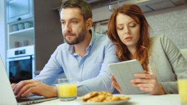 Genç çift mutfakta siting. Tablet kocası dizüstü kullanarak süre kullanan genç güzel kadın. Kapalı.