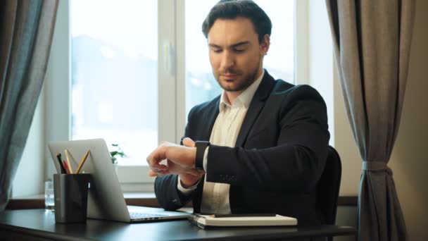 Молодий серйозний бізнесмен у костюмі важко працює з ноутбуком і час від часу дивиться на смарт-годинник на офісному фоні з вікном . — стокове відео